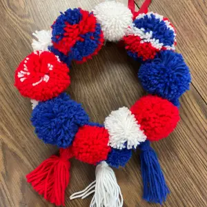 Yarn pom-pom wreath in red, white, and blue on a wooden surface, links to more information